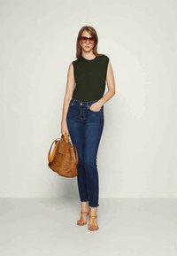 Top sem mangas verde oliva, jeans skinny azul escuro, saltos laranja de tiras e uma bolsa de ráfia castanha com detalhes em tom de pele.