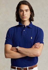 Sinine lühikeste varrukatega polo särk, millel on krae ja kaks nuppu, esiküljel vasakul rinnal väike kollane hobuse logo. Kandmine pruun vööga.