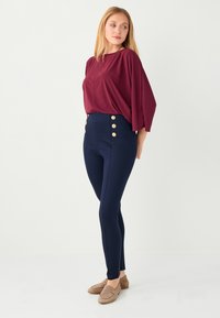 Blouse bordeaux à manches chauve-souris associée à un pantalon taille haute bleu marine avec des accents de boutons dorés sur le côté. Matière douce et extensible.