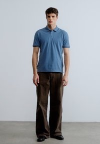 Gaiši zils polo krekls ar īsām piedurknēm, kombinēts ar brūnām platām biksēm. Krekls ir ar standarta apkakli un smalku logotipu.