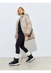 Doudoune beige avec capuche et accents tricotés, associée à un legging noir et des bottes d'hiver noires avec des manchons blancs.