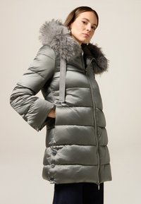 Giubbotto trapuntato grigio con cappuccio in pelliccia sintetica, chiusura con zip e tasche laterali. Il tessuto ha una texture liscia e lucida con elementi di design trapuntato.