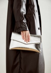Stříbrná metalická kabelka typu clutch se sklápěcím víkem, elegantní design, hladká textura, držená v ruce, zkombinovaná s černým kabátem.
