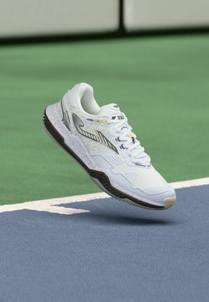 Chaussure de sport blanche avec des accents noirs et jaunes flottant au-dessus de la surface bleue et verte du court de tennis près des lignes du court.