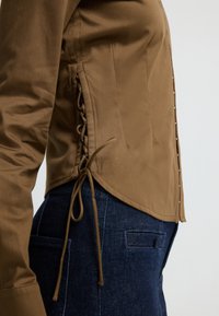 Brown long-sleeve shirt with side lace-up detail paired with dark blue denim jeans, shown from the waist to mid-torso side view.