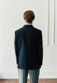 Tumesinine bleiser, millel on struktureeritud disain, servadega krae ja sile tekstuur, kombineeritud helesiniste pükstega. Näidatud tagantvaade.