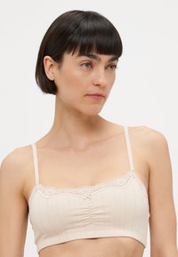 Lys beige bralette med mykt stoff, blondekant langs utringningen, justerbare stropper og subtile vertikale ribbestrukturdetaljer.