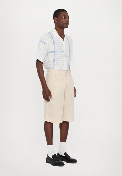Chemise à manches courtes à carreaux blancs et bleus, associée à un short plissé beige clair. Le modèle porte des chaussures noires et des chaussettes blanches.