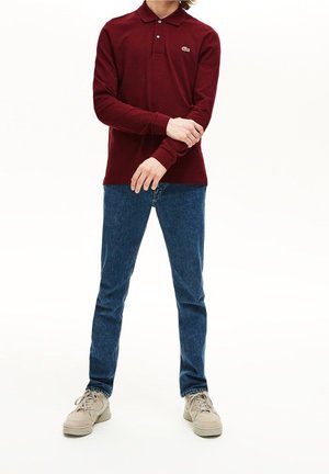 Persona che indossa una polo bordeaux a maniche lunghe, jeans blu e sneakers beige, in piedi con le braccia incrociate ai polsi.