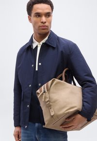 Νεαρός άνδρας με μπλε μπουφάν και τζιν κρατά μια μεγάλη μπεζ δερμάτινη τσάντα με καφέ λαβές, κοιτώντας προς το πλάι σε απλό φόντο.