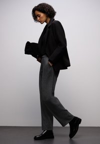 Zwarte op maat gemaakte blazer, zwarte top, grijze gestructureerde wijde broek en zwarte enkellaarsjes. Model poseert met een serieuze uitdrukking.