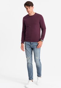 Sweat-shirt bordeaux à col rond, associé à un jean déchiré bleu clair. Le mannequin porte des baskets blanches et une montre en argent.