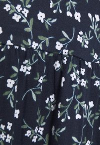 Mörk tyg med små vita blommor och gröna blad i ett spritt blommönster, med detaljer av rynkade sömmar.