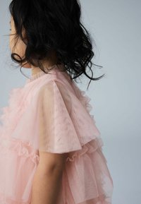 Roza tila obleka z večplastnimi volani, prosojnimi rokavi in teksturiranim zgornjim delom. Mehka, lahka tkanina z nežnim, zračnim videzom.