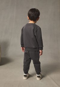 Malo dijete kratke tamne kose stoji okrenuto leđima, nosi tamnosivu majicu s dugim rukavima, odgovarajuće hlače, bijele čarape i bež tenisice.