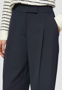Pantalons plissés bleu marine en tissu lisse, avec une coupe ajustée et des poches latérales, associés à un pull en maille rayé.