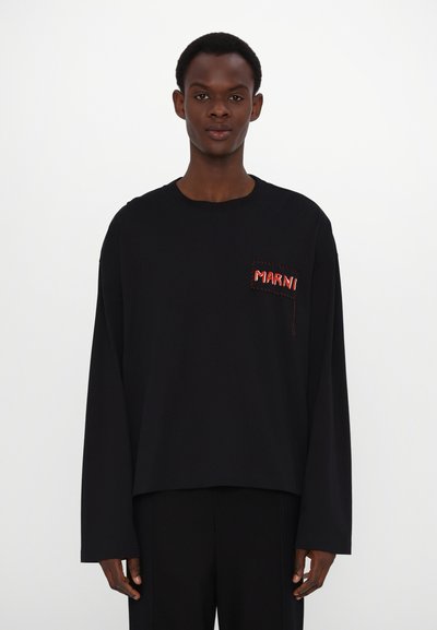 T-shirt noir à manches longues en coton, avec un logo rouge brodé sur une poche patch. Coupe loose, encolure ronde et coutures d'épaules tombantes.
