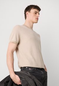 Beige, rövid ujjú pulóver texturált kötésből, kerek nyakkivágással és bordás mandzsettával, sötét farmernadrággal és dzsekivel párosítva.
