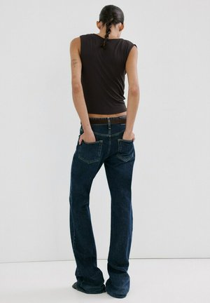 Ženska z pletenicami, oblečena v črno majico brez rokavov in temno modre kavbojke, z rokami v zadnjih žepih, stoji ob belem ozadju.