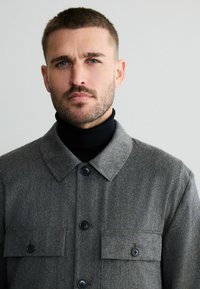 Muž s krátkymi vlasmi a bradou, ktorý má na sebe šedý zapínací kabát cez čierny rolák, pozerá spredu s neutrálnym výrazom na jednoduchom pozadí.