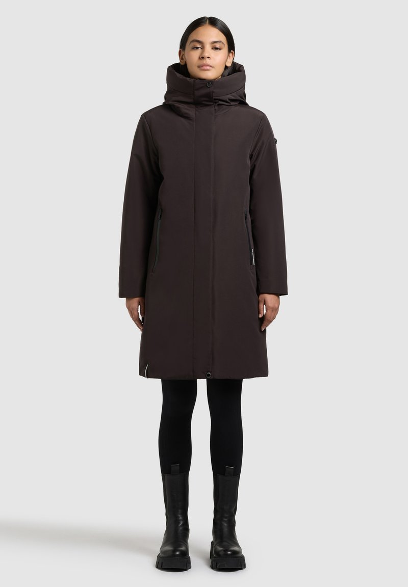 Long dark brown coat with a high collar, zipper closures, and side pockets. Worn over black leggings and sturdy black boots.