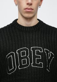 Pulover negru tricotat cu textură rib de culoare neagră, având cuvântul "OBEY" scris cu litere mari albe pe piept. Guler rotund, croială casual.