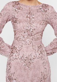 Vestido rosa claro con mangas largas que presenta intrincados bordados florales y aplicaciones de cuentas. La tela tiene una textura suave con acentos brillantes.