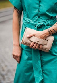 Bežová kabelka typu clutch držená v ruce, s obdélníkovým tvarem, texturovanou kůží a výraznou logem. Nositelná s zelenými šaty.