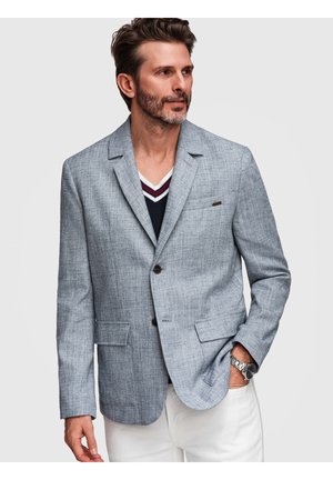 Homme aux cheveux bruns courts et à la barbe, portant un blazer gris, un pull col en V bleu marine, un pantalon blanc et une montre-bracelet, regardant vers la droite.