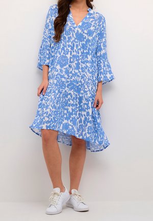 Kvinde iført en blå og hvid knælængde kjole med blomsterprint, klokkeærmer og hvide sneakers, stående mod en ensfarvet baggrund.