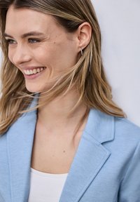 Jeune femme aux cheveux châtain clair souriante, portant de petites boucles d'oreilles en argent, un haut blanc et un blazer bleu clair sur un fond uni.