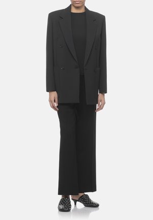 Persona che indossa un blazer nero doppiopetto oversize, pantaloni neri a gamba larga, top nero e mules nere con borchie e tacco.