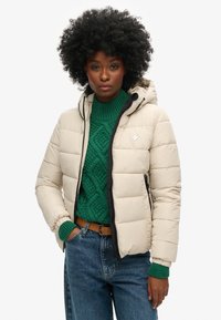 Beige vadderad jacka med huva, med svarta kantband och dragkedjedetaljer. Bärs över en grön texturerad stickad tröja och blå jeans.