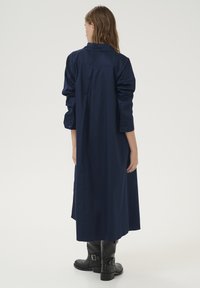Rochie din bumbac, de culoare bleumarin, cu mânecă lungă, având un pliu la spate și manșete cu nasturi, asortată cu cizme negre până la genunchi.