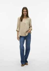 Beige košile s dlouhým rukávem a výstřihem do V, s předním zapínáním na knoflíky, spárovaná s volnými modrými džíny a hnědými botami.