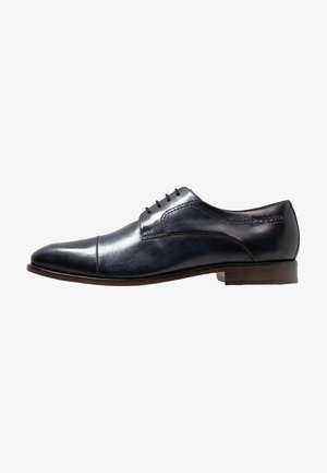 Chaussure oxford en cuir noir avec une finition lisse, bout rond, cinq œillets et un petit talon marron. Présente des coutures discrètes et un logo.