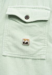 Τσέπη πουκαμίσου από ανοιχτό πράσινο κορδόνι με καφέ κουμπί και μικρό ορθογώνιο μπάλωμα με κίτρινα και μαύρα κυματιστά σχέδια.