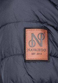 Marinblått puffjacka med en broderad läderlapp som visar "NAVAHOO EST 2014" och logotypdesign. Slät tygtextur, isolerad.