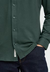 Donkergroen langrechtshirt met een knoopsluiting aan de voorkant, gestructuurde stof, de zoom is licht gebogen, gecombineerd met denim jeans. Label zichtbaar aan de zijkant.