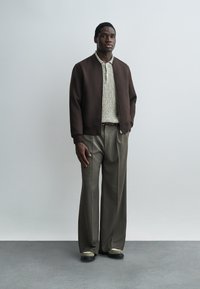 Junger Mann, der eine braune Bomberjacke, einen grauen Strickpullover, weit geschnittene graue Hosen und schwarz-weiße Schuhe trägt, steht vor einer schlichten Wand.