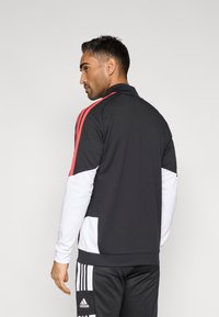 Fekete sportzubbony piros részletekkel, fehér ujjakkal, cipzáras gallérral és logó díszítéssel. Simább textúra, kontrasztos színblokkokkal.