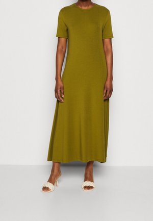 Osoba stojąca w oliwkowo-zielonej sukience za kolana z krótkim rękawem, nosząca białe sandały na obcasie z odkrytymi palcami, na jednolitym tle.