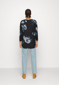 Schwarzes Langarmoberteil mit Blumenmuster in Blau- und Grüntönen, kombiniert mit hellblauen Skinny-Jeans und beigen Absatzmules.