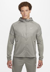 Chaqueta Nike de color gris claro con cierre de cremallera frontal, capucha, tejido texturizado y paneles de malla. Cuenta con bolsillos laterales y un pequeño logo en el pecho.