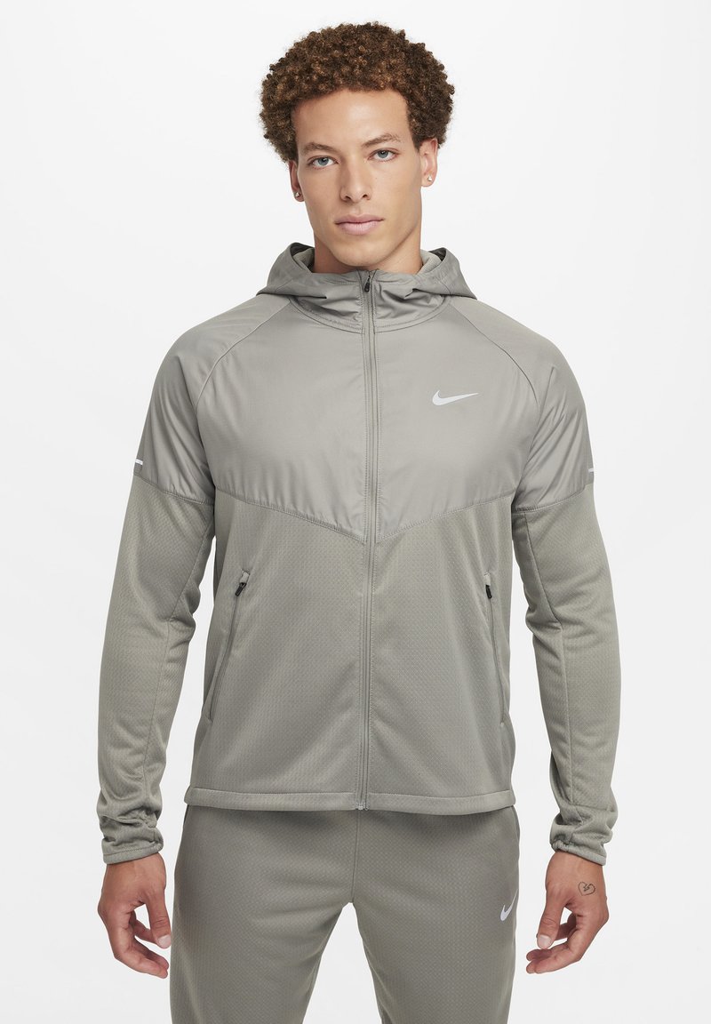 Chaqueta Nike de color gris claro con cierre de cremallera frontal, capucha, tejido texturizado y paneles de malla. Cuenta con bolsillos laterales y un pequeño logo en el pecho.