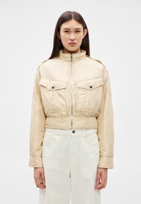 Sieviete ar taisniem tumšiem matiem, valkājot bēša krāsas īsu žaketi ar atloku kabatām un baltas augsta vidukļa bikses uz vienkārša fona.