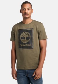 T-shirt di cotone verde oliva con collo rotondo; presenta un logo grafico di un albero nero e il testo "Timberland" all'interno di un riquadro rettangolare.