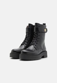 Botas de tobillo de cuero negro con punta redondeada, cordones en la parte frontal, suela gruesa y una correa decorativa con un acento dorado.
