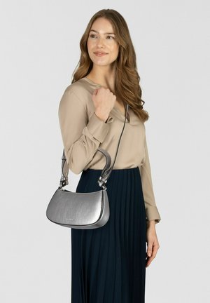 Donna con camicetta beige e gonna blu navy che porta una borsa a tracolla grigio metallico con una tracolla corta e una lunga, che sorride e guarda di lato.