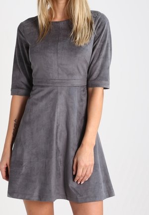 Kvinde iført en kortærmet, knælængde grå ruskindskjole med en figursyet talje mod en ensartet hvid baggrund.